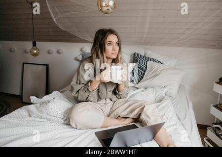 Nachdenkliche Frau mit Laptop, die Kaffee trinkt, während sie zu Hause auf dem Bett sitzt Stockfoto