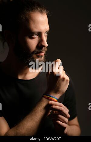 Porträt eines bärtigen jungen Mannes mit Regenbogenarmband vor dunklem Hintergrund Stockfoto