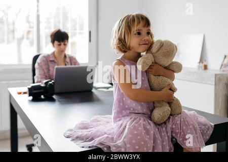 Mutter arbeitet zu Hause, Tochter sitzt mit Teddybär auf dem Tisch Stockfoto