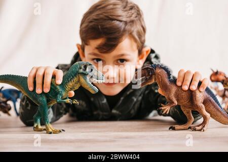 Lächelnder kleiner Junge, der auf dem Boden liegt und mit Spielzeug-Dinosauriern spielt Stockfoto