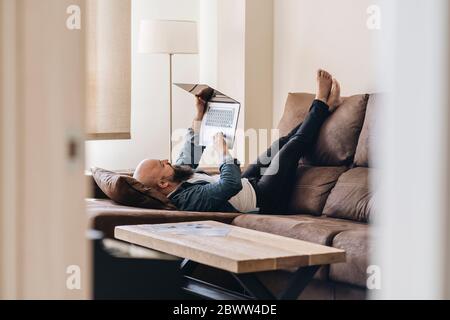 Gelangweilter Mann mit Laptop, während er im Wohnzimmer zu Hause auf dem Sofa lag Stockfoto