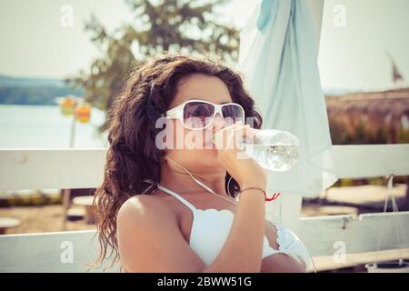 Im Sommer können Sie sich im Freien mit Feuchtigkeit versorgen. Gut aussehende Mixed Race Frau in Sonnenbrille mit Afro Haarschnitt in weißen Badeanzug, Sonne genießen, dri Stockfoto