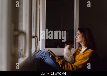 Lächelnde rothaarige Frau, die am offenen Fenster sitzt und ihre Katze aus der Ferne betrachtet Stockfoto