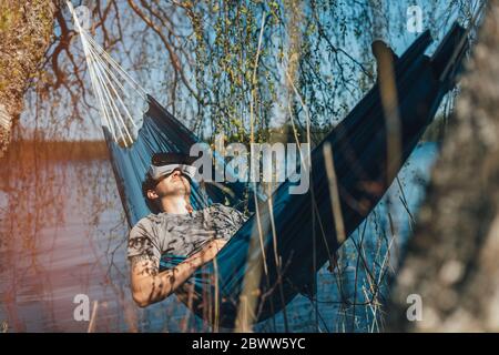 Junger Mann, der in der Hängematte entspannt und VR-Brillen verwendet Stockfoto