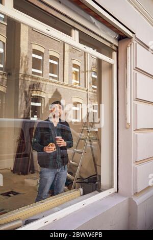 Mann, der die Lage des Ladens renoviert, Kaffee trinkt und aus dem Fenster schaut Stockfoto