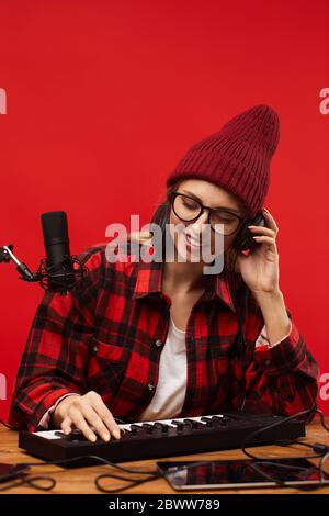 Junge lächelnde Frau in Kopfhörer sitzen am Tisch und mit Musiktastatur sie Aufnahme des Songs Stockfoto