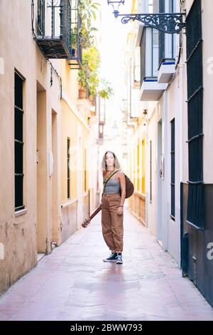 Porträt einer jungen Frau mit Gitarre, die in einer engen Straße inmitten von Gebäuden in Santa Cruz, Sevilla, Spanien, steht Stockfoto