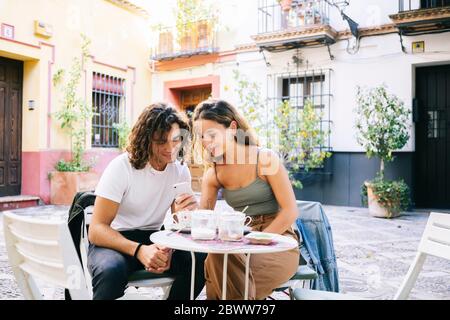 Junge Frau, die ihrem Freund Handy zeigt, während sie im Straßencafé in Santa Cruz, Sevilla, Spanien sitzt Stockfoto