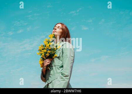 Porträt einer rothaarigen jungen Frau mit geschlossenen Augen, die gegen den Himmel steht und einen Strauß gelber Blumen hält Stockfoto