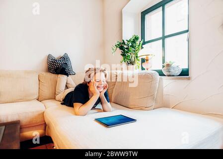 Lächelnder Junge, der mit einem Tablet im Wohnzimmer auf der Couch liegt Stockfoto