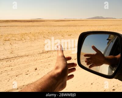 Spiegelung im Rückspiegel einer Menschenhand vor dem Fenster während der Fahrt in der Wüste, Sossusvlei, Namibia. Stockfoto