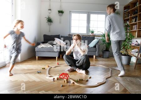 Gestresste Mutter sitzt mitten in Spielzeug, während Kinder um sie herum laufen Stockfoto