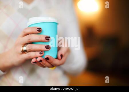 Ernte anonyme Frau in warmen Pullover mit dunkler Maniküre hält Einweg Tasse Kaffee während der Ruhepause am Holztisch Stockfoto