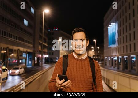 Porträt eines lächelnden Geschäftsmannes mit Smartphone bei Nacht, Frankfurt, Deutschland Stockfoto