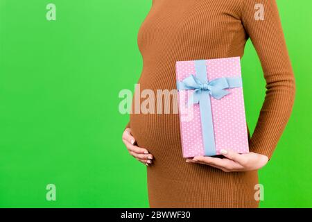 Cropped Bild der schwangeren Frau in braunem Kleid mit rosa gepunkteten Geschenk-Box auf grünem Hintergrund. Warten auf ein Baby Mädchen. Kopierbereich. Stockfoto