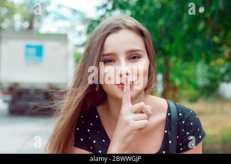 Shh Frau mit breiten Augen bitten um Stille oder Geheimhaltung mit Finger auf den Lippen Hush Hand Geste Stadtpark Outdoor Hintergrund hübsches Mädchen, das Finger auf li Stockfoto