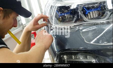 Nahaufnahme des PPF-Installationsprozesses an einem Scheinwerfer und einer Motorhaube. PPF ist eine Lackschutzfolie, die Farbe vor Kratzern und Steinschlag schützt. Stockfoto