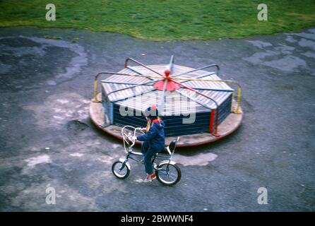 Ein 7-jähriges Mädchen, das blaue Jeans, einen blauen Wollmütze mit rotem Bommel und einer blauen Jacke trägt und mit ihrem Raleigh Tomahawk Fahrrad um einen Spielplatz-Kreisverkehr in Hatch End, Middlesex, Großbritannien, fährt Stockfoto