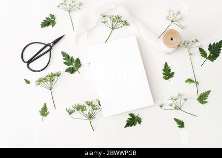 Hochzeit, Geburtstag Desktop Mock-up-Szene. Leere Grußkarte, schwarze Vintage-Schere, Seidenband und KuhPetersilie Blätter und Blumen. Weißer Tisch Stockfoto