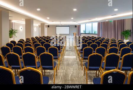 Vorderansicht des luxuriösen Auditoriums mit leerem weißen Bildschirm. Moderner Konferenzsaal mit Reihen von blauen und goldenen Stühlen Stockfoto