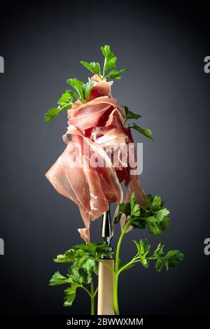 Dünnes geschnittenes Prosciutto mit Petersilie auf einer Gabel. Traditionelle italienische Küche. Schwarzer Hintergrund. Stockfoto