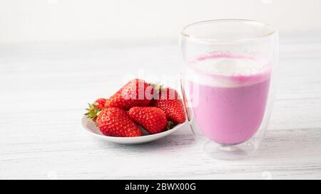 Wenige reife Erdbeeren und rosa Matcha Latte mit Milch in doppelwandigem Glas auf weißem kleinen Teller auf Holztisch Stockfoto