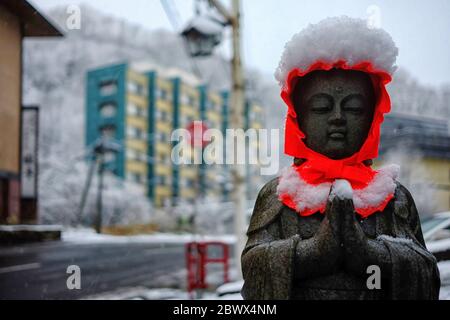 Altes Stein-Buddha-Bild mit roter Kapuze im fallenden Schnee in Noboribetsu Onsen, wo ist berühmte Touristenattraktion in Hokkaido, Japan. Stockfoto