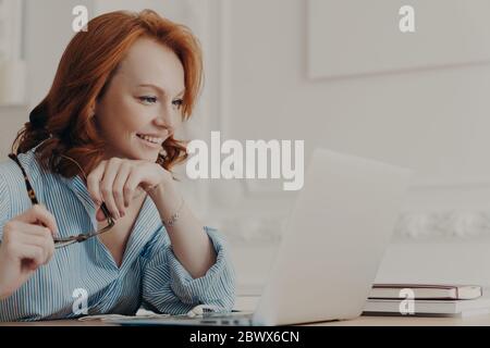 Horizontale Aufnahme von Rotschopf Europäische Frau posiert im Home Office, auf Laptop-Bildschirm fokussiert, hält Brillen, sieht Video auf Computer, genießt gute neue Stockfoto