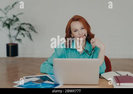 Schöne Rotschopf Frau arbeitet entfernt von zu Hause, sitzt vor Laptop-Computer beendet die Arbeit an Projekt denkt über das Hinzufügen von interessanten conten Stockfoto
