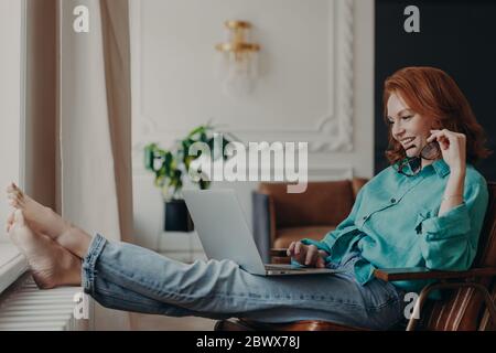 Entspannte Studentin mit Ingwerhaar in Hemd und Jeans, hält Laptop auf den Knien, durchstöbert notwendige Informationen, bereitet sich auf das Bestehen der Prüfungen vor. Stockfoto