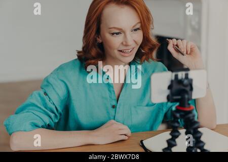 Junge professionelle Frau Vlogger oder Blogger posiert vor der Smartphone-Kamera, gibt Ratschläge, wie man eigenes Geschäft zu starten, sitzt am Desktop, hält Stift. P Stockfoto