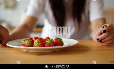 Frau, die Erdbeeren auf den Teller legt. Nahaufnahme von Frauenhänden, die frische Beeren auf den weißen Teller auf den Küchentisch legen. Stockfoto