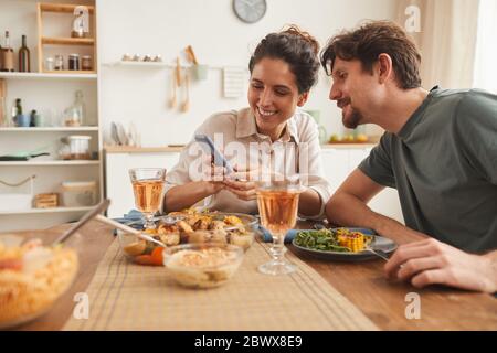 Junge lächelnde Frau zeigt ihrem Freund während des Abendessens in der Küche zu Hause einige Bilder auf ihrem Handy Stockfoto