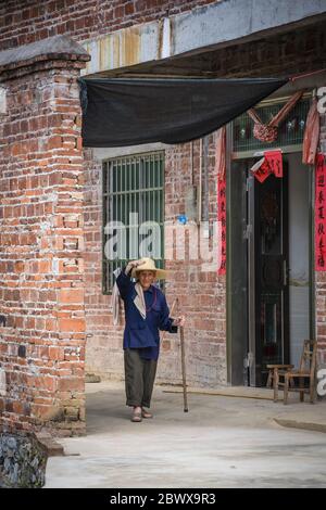 Yangshuo, China - August 2019 : Alte chinesische Dame mit Strohhut und Holzstab zu Fuß auf dem Hof vor ihrem Dorf zu Hause in ru Stockfoto