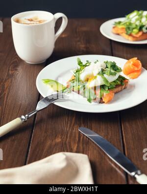 Frühstück mit Kaffee und Toast mit Avocado, Lachs Fisch, pochiertes Ei, Rucola Salate auf einem weißen Teller. Gesundes vegetarisches Gericht. Stockfoto