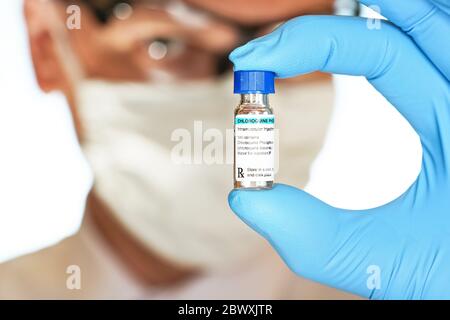 Hand in blauen medizinischen Handschuhen hält kleine Fläschchen mit Chloroquine Phosphat (generischer Name, eigenes Design-Label), verschwommenes Gesicht in Baumwollmaske und Brille BA Stockfoto