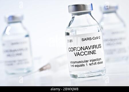 Coronavirus Covid-19 Impfstoff-Konzept - Glasfläschchen mit Metallkappen auf weißem Tisch, verschwommene hypodermische Spritzennadel und mehr Flaschen im Hintergrund Stockfoto
