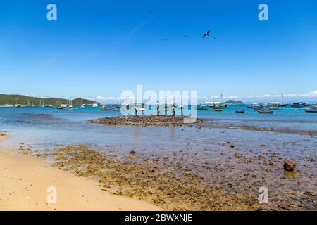 BUZIOS, BRASILIEN. Panoramablick auf die Bucht von Armacao und die Bronzeskulptur drei Fischer. Viele und bunte Boote ankern im Meer. Stockfoto