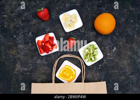 Kreative flache Lage von ganzen und geschnittenen Früchten auf kleinen Tellern fallen in eine Papier-Einkaufstasche auf einem grunge dunklen Hintergrund. Gesunde Ernährung und Bio Stockfoto