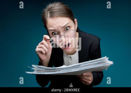 Eine junge Frau legt ihr eine Brille auf die Nase und schaut mit verärgerlicher Überraschung auf die Dokumente. Direktor oder Lehrer unzufrieden mit Papierkram oder Fehler in ihnen Stockfoto