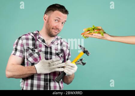 Ein schmutziger harter Arbeiter mit Werkzeugen in den Händen zieht einen Schutzhandschuh aus seiner Hand und lächelt schlaff, weil er ein Sandwich hält. Snack danach Stockfoto