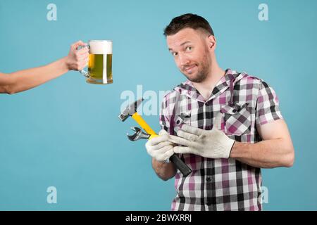 Ein schmutziger harter Arbeiter mit Werkzeugen in den Händen zieht einen Schutzhandschuh aus seiner Hand und lächelt schlaff, weil jemand ein Bier hält. Ruhe danach aus Stockfoto