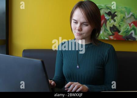 Ein Mädchen mit einem nachdenklichen, angespannten Gesichtsausdruck sitzt auf einem braunen Sofa an einem Laptop an einem Tisch in einem Café an einer gelben Wand. Er schaut mit verengten Augen auf den Computerbildschirm. Stockfoto