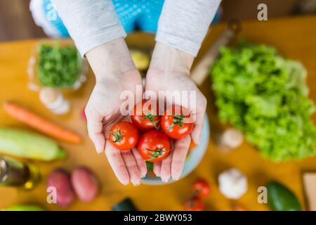 Hohe Winkel Ansicht der Frau Hände halten Kirschtomaten gegen Tisch voller verschiedener Gemüse in der Küche. Vegetarisches Essen Konzept. Stockfoto