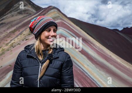 Schöne blonde Frau im Regenbogenberg in Peru mit allen Farben des Berges im Hintergrund. Stockfoto