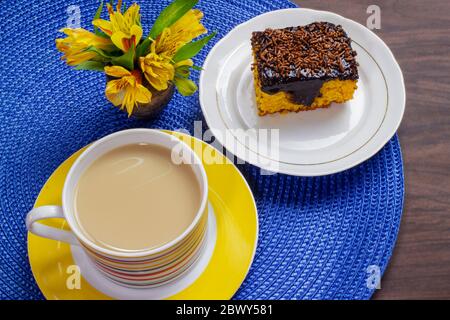 Karottenkuchen mit Schokoladenglasur und Tasse Milch mit Kaffee auf Holztisch mit einer gelben Blume dekoriert. Stockfoto