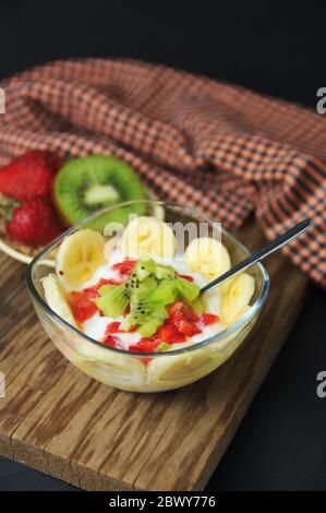 Schüssel mit einem gesunden Frühstück oder Snack aus Joghurt und Obst wie Kiwi, Bananen, Erdbeeren, auf einem Holztisch und schwarzem Hintergrund mit sp Stockfoto
