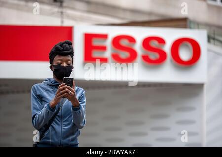 Frau, die ein Kopftuch und eine Gesichtsmaske trägt, mit einem Mobiltelefon vor einem ESSO-Garagenschild. London, England, Großbritannien Stockfoto