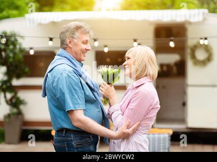Wir feiern das Jubiläum. Reife Frau, die Blumen hält, die ihr fürsorglicher Ehemann auf dem Campingplatz gegeben hat Stockfoto