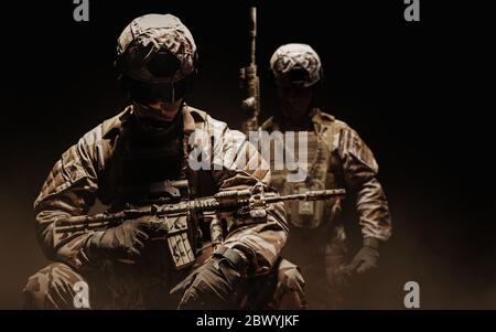 Foto von einem voll ausgestatteten Wüste Soldaten in getarnten Rüstung sitzen und stehen mit Gewehr auf dunklen nebligen Hintergrund. Stockfoto